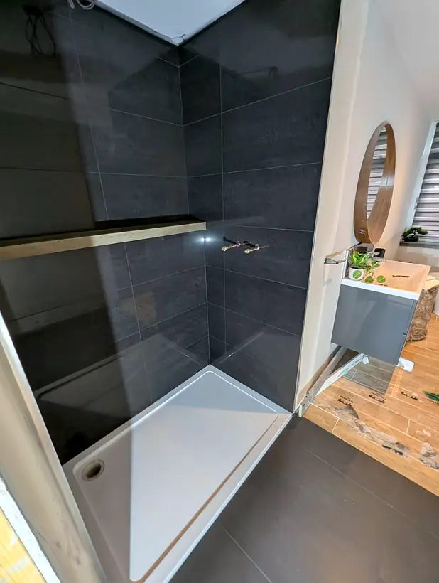 Walk in shower with large dark tiles and an oak shelf